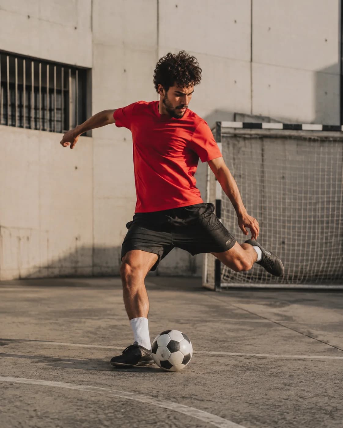 A player in a red jersey winding up to strike a football on an urban court