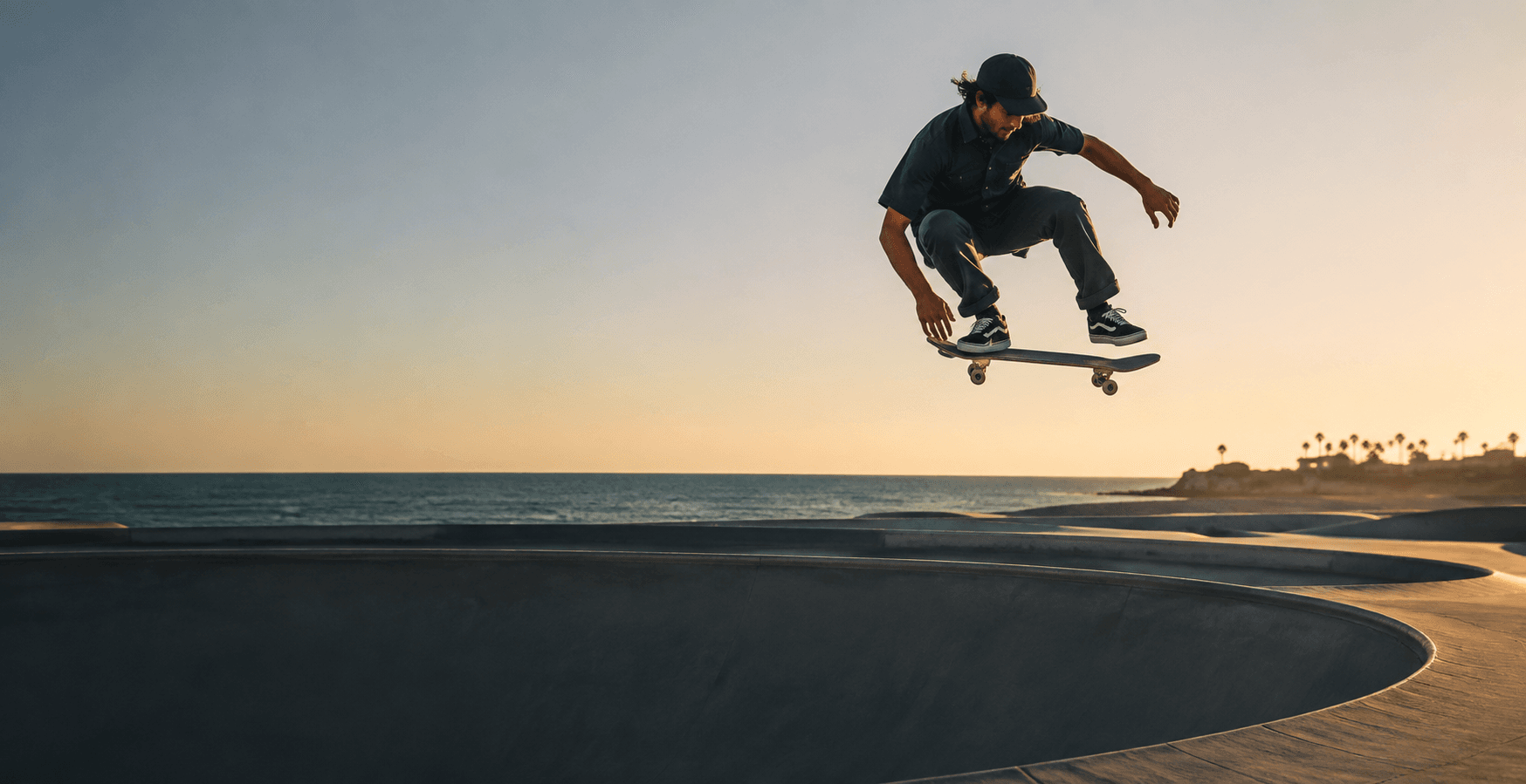 A skateboarder suspended above a coastal skate bowl at sunset